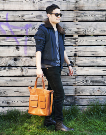 Homme tenant un sac à dos en cuir orange vif, vue de face, devant un mur en bois gris
