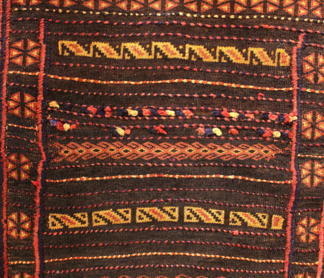 Rust-red textile with geometric striped patterns in gold and cream, featuring traditional woven design with borders