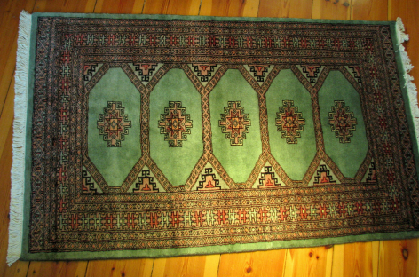 Green and gold patterned textile rug with ornate borders, displayed flat on wooden floor, overhead view