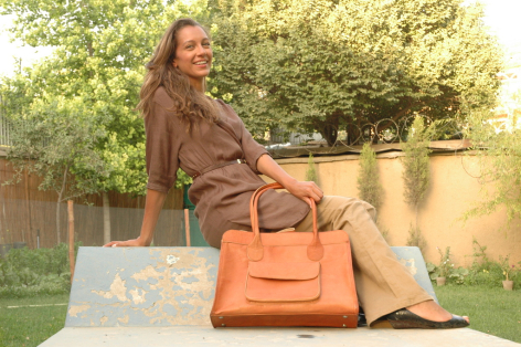 Woman sitting outdoors holding a tan leather handbag, wearing brown clothing against a green garden backdrop