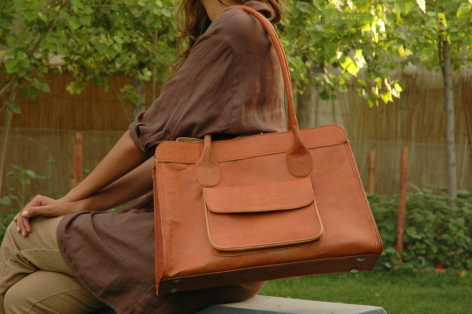 Tan leather tote bag with structured silhouette and front patch pockets, displayed open against green foliage background