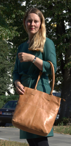 Woman wearing dark teal jacket holding tan leather shoulder bag with front view against green foliage background