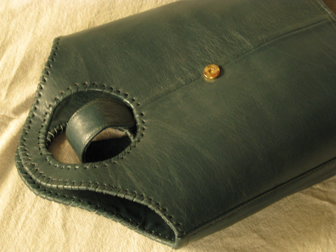 Sac en cuir vert foncé plié, vue de dessus montrant un détail de couture artisanale et une petite perle dorée