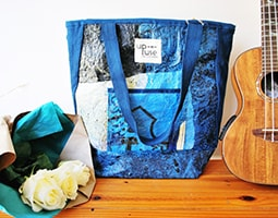 Blue patterned tote bag displayed next to wooden guitar, white flowers, and teal fabric on wooden surface