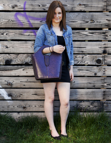 Woman wearing blue cardigan and holding deep purple leather crossbody bag against weathered wooden wall background