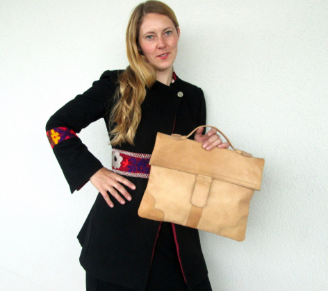 Woman wearing black jacket holding tan leather bag with front flap pocket, angled toward camera