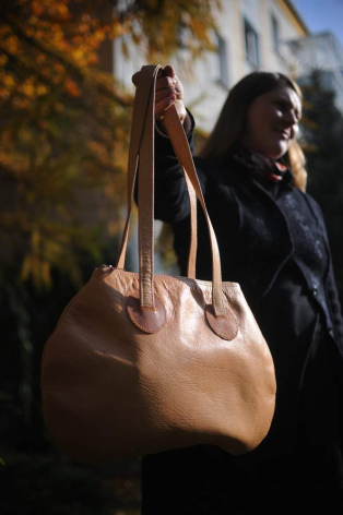 Tan leather tote bag with dual handles held by person wearing dark clothing, autumn foliage visible in background