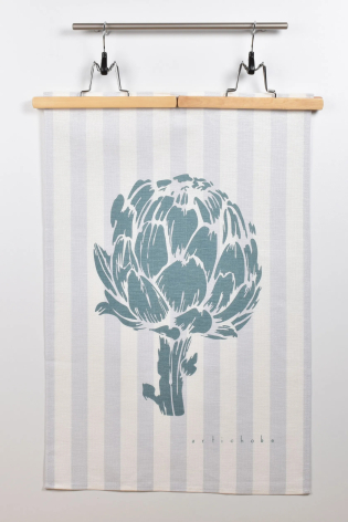 Cream linen tea towel with blue artichoke illustration, hung on wooden dowel with metal clips, front view