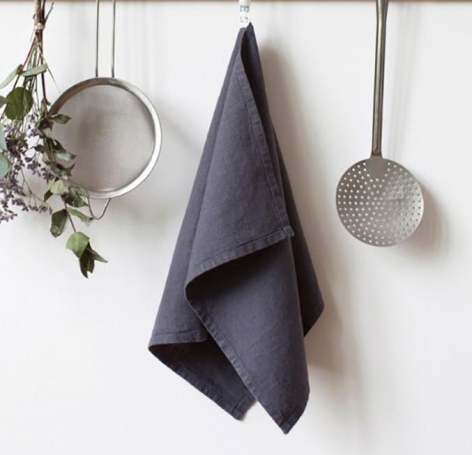 Dark gray linen kitchen towel hanging centrally between two hanging sieves and dried herbs against white wall
