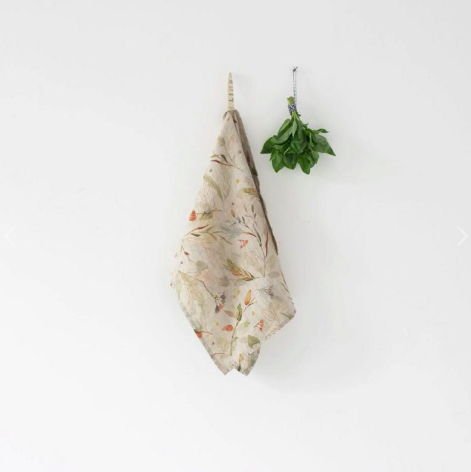 Beige linen kitchen towel with subtle pattern hanging from string beside dried green herbs against white background
