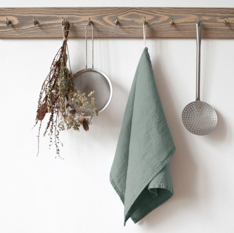 Wooden wall rack with dried botanicals, metal sieves, and sage green linen against white wall
