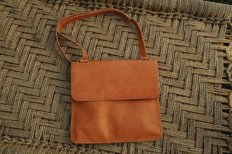 Tan leather shoulder bag with flap closure, displayed flat on woven geometric patterned textile background