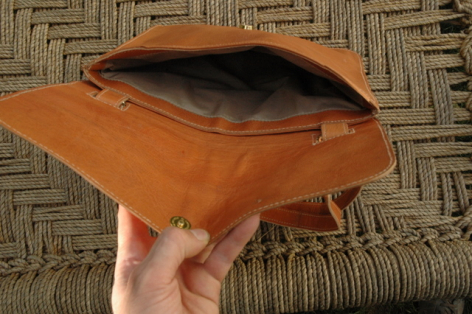 Tan leather clutch bag held open in hands, showing interior compartment against woven textile background