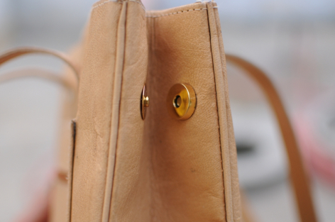 Tan leather bag shown from side view with brass circular clasp detail and natural handle visible
