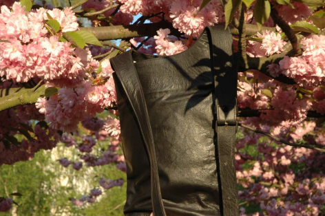 Olive green canvas crossbody bag with black leather accents, displayed against pink and white flowering plants