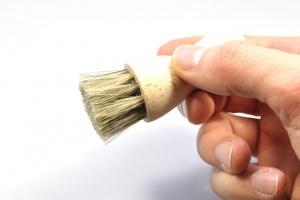 Person's hand holding a round wooden brush with natural bristles, shown in profile against white background