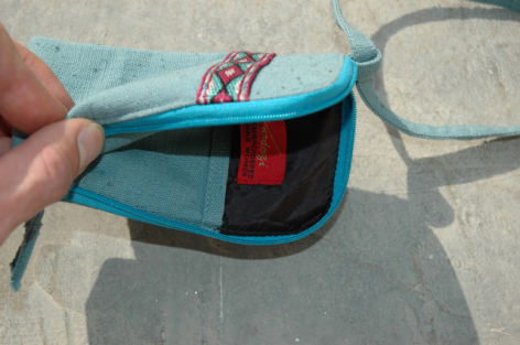 Bright blue fabric waist bag with red zippered pocket, held open in hands against concrete surface